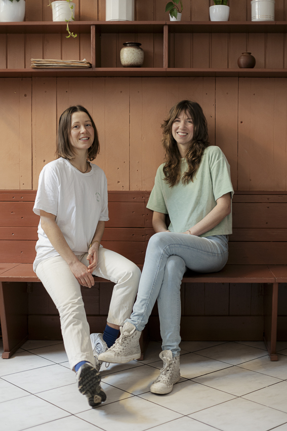 Die Unternehmerinnen Constanze Karácsonyi und Theresa Rauch in pastellfarbener Kleidung vor einer braunen Wand.