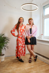 Laura und Veronika in hellen Farben, neben ihnen eine Pflanze am Boden.